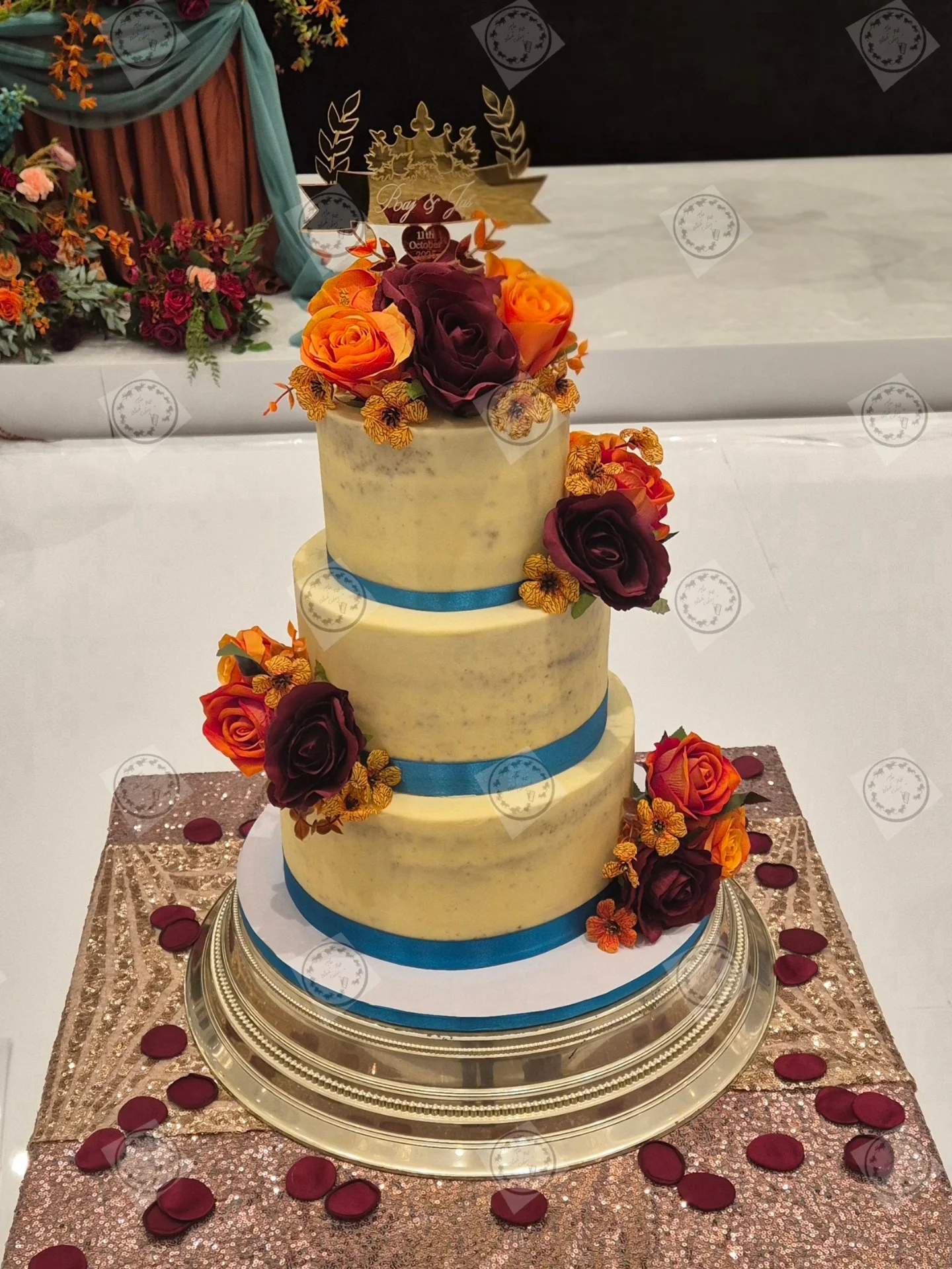 Three-tier semi-naked wedding cake with burgundy and burnt-orange silk roses, gold ribbon detailing and a personalised gold cake topper